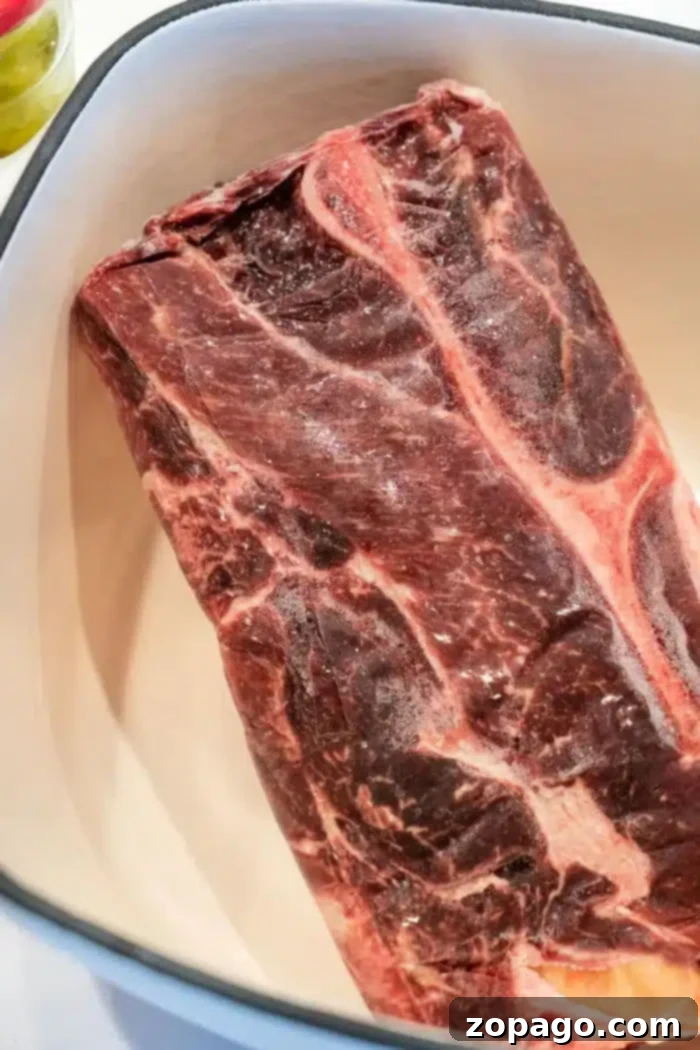 A raw beef chuck roast in a white oblong dish, ready to become tender shredded beef from frozen. The meat has visible marbling and fat along the edges.