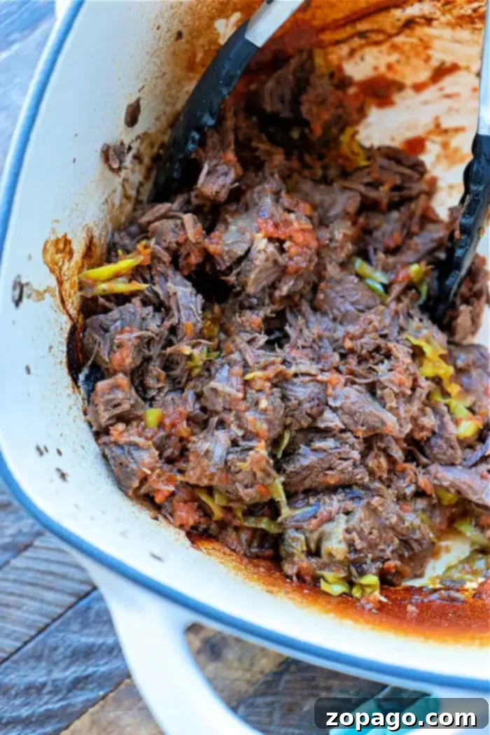 A white pot filled with shredded beef, made from frozen, includes visible chunks of vegetables like tomatoes and green peppers. Black tongs rest inside the pot, and sauce residue clings to the sides. The pot is set upon a wooden surface.