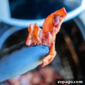 A gloved hand proudly lifts a crispy bacon strip, freshly cooked from a frozen state in the air fryer.