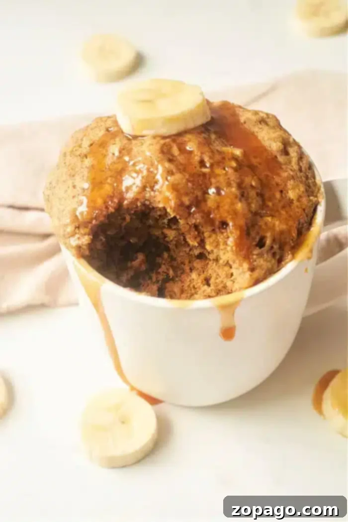 A Banana Bread in a Mug creation, topped with a banana slice and drizzled with syrup. The cake has a bite taken out of it, revealing its moist texture. Additional banana slices are artfully scattered around the mug on a light surface, enhancing its delicious appeal.