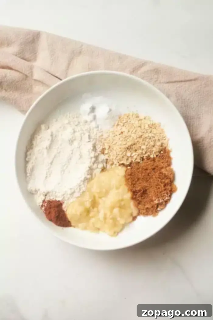 A white bowl holds the makings of banana bread in a mug: flour, oats, brown sugar, mashed banana, cinnamon, and a dash of baking soda. A beige cloth subtly peeks into view on the pristine white surface.