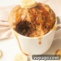 Banana Bread in a Mug: A mouthwatering banana mug cake with a bite taken, topped with fresh banana slices and drizzled syrup, all on a pristine white surface.