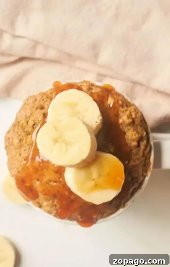 Microwave Banana Bread Bliss 6 A banana bread mug cake topped with sliced bananas and drizzled with syrup is placed against a soft pink background. Some banana slices are also visible on the surface near the mug.