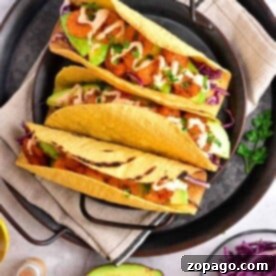Three tacos with chicken, avocado, cabbage, and sauce sit invitingly on a plate. Beside them is a fresh avocado half and vibrant purple cabbage. These delicious tacos feature perfectly crispy taco shells made in an air fryer for the ultimate crunch experience.