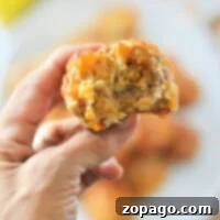 Savory Bisquick Sausage Bites 7 A hand holds a bitten savory Bisquick sausage balls, with a blurred background showcasing similar muffins on a plate.