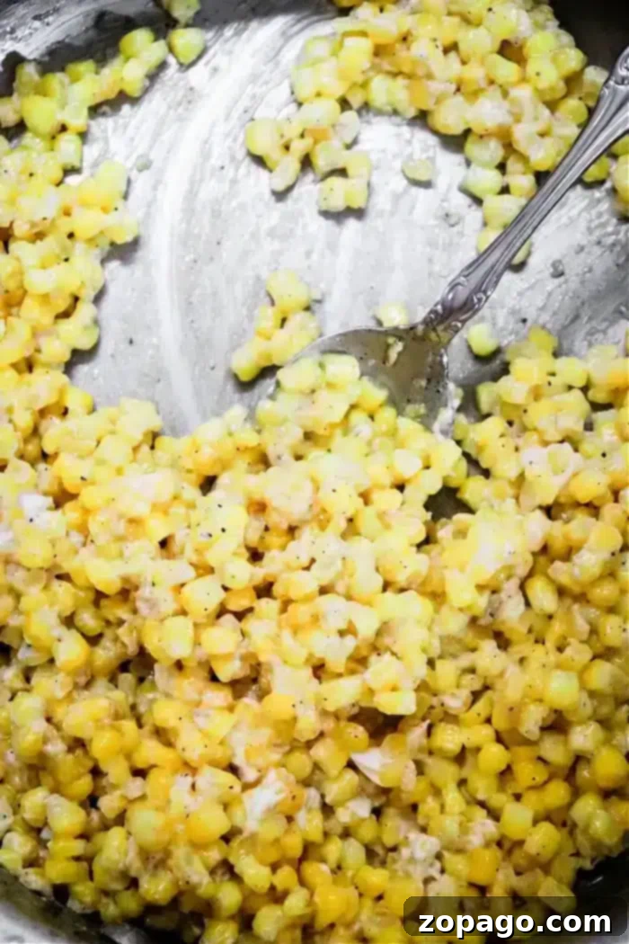 Golden Honey Butter Skillet Corn 7 A close-up of a metal dish containing honey butter skillet creamed corn. The yellow kernels are mixed with a creamy sauce, accented by black pepper, while a metal spoon rests in the dish, partially immersed in the corn mixture.