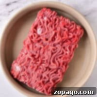 Frozen ground beef rests in a round beige dish on a marble surface, ready for you to thaw ground beef in the microwave quickly and easily.
