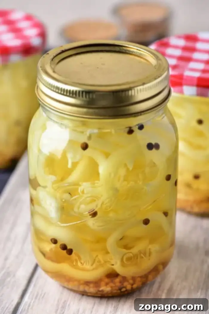 Crisp Golden Banana Pepper Rings 2 A mason jar filled with vibrant pickled banana pepper slices and mustard seeds, topped with a golden lid, showcases the delicious result. In the background, other jars with checkered lids are nestled on a rustic wooden surface, hinting at a broader world of homemade preserves.