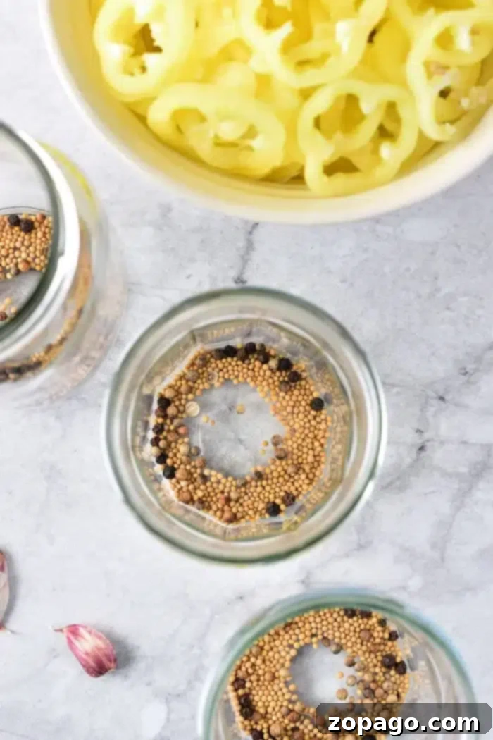 Crisp Golden Banana Pepper Rings 3 Three empty glass jars with mustard seeds and peppercorns sit on a polished marble surface, poised for preserving a batch of homemade pickled vegetables. A vibrant bowl of sliced yellow peppers and a solitary clove of garlic nearby set the scene for an upcoming culinary creation.