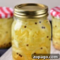 Crisp Golden Banana Pepper Rings 5 A jar of pickled banana peppers takes center stage, showcasing a metal lid and visible peppercorns. In the blurred background, two more jars with checkered lids hint at an assortment of pickled fruits and vegetables, all elegantly displayed on a wooden surface.