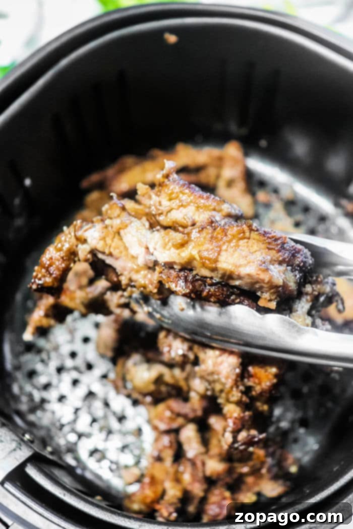 Air Fryer Mongolian Beef 4 Close-up of tongs gripping perfectly cooked beef strips in an air fryer basket, showcasing the crispy delight.