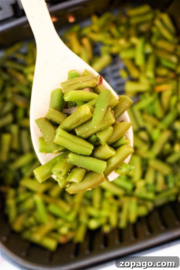 Crispy Air Fryer Frozen Green Beans 7 Close-up of a spoon holding air fried green beans over a pan filled with more green beans, showcasing the vibrant freshness often missing in air fryer frozen green beans.