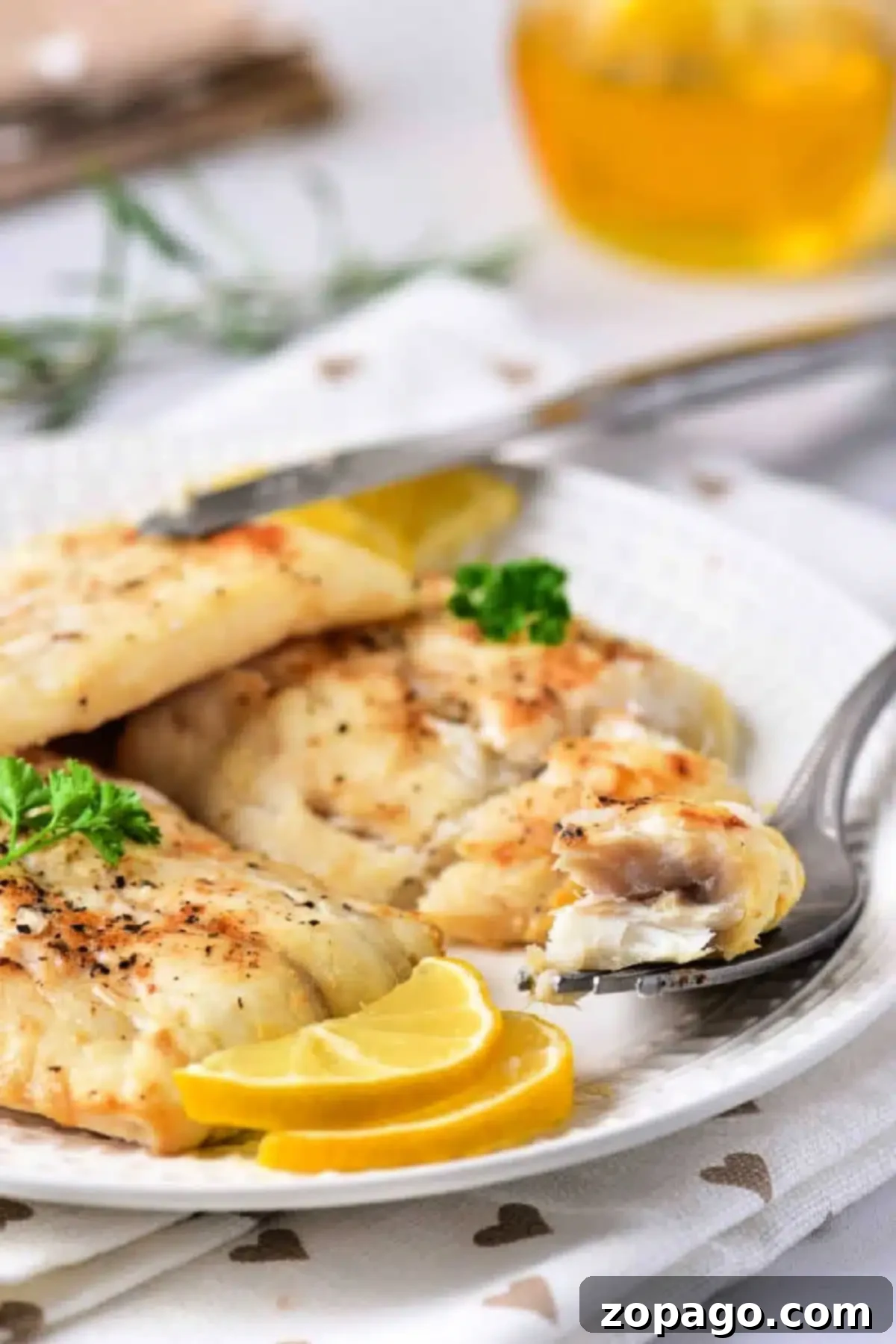 Crispy Air Fryer Cod 2 A white plate features air-fried fish fillets garnished with lemon slices and a sprig of parsley. A fork holds a piece of the flaky delight, while a blurred glass with a yellow drink sits in the background. The tablecloth is white with heart patterns, emphasizing this delicious twist on frozen cod.