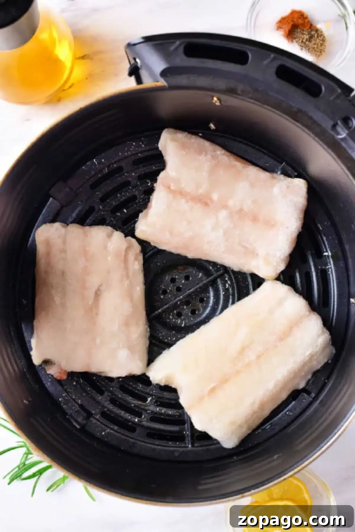 Crispy Air Fryer Cod 5 Three frozen cod fillets are placed in an air fryer basket. Nearby, there is a small bowl of spices, a glass bottle of oil, rosemary sprigs, and lemon slices on a light-colored surface.