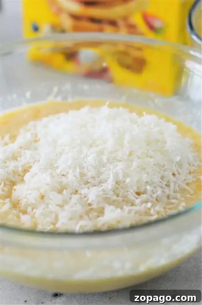 Easy Crustless Coconut Bisquick Impossible Pie 5 A glass mixing bowl holds the prepared Bisquick Impossible Coconut Pie batter, generously topped with shredded coconut before baking.