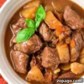 A bowl of savory beef stew, featuring tender chunks of meat, potatoes, carrots, and a fresh basil garnish, rests invitingly on a red cloth background. This delicious beef stew recipe comes together perfectly without wine.