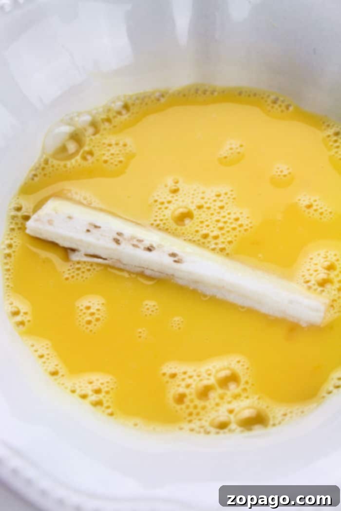 Eggplant stick being dipped into an egg mixture in a white bowl, preparing for its transformation into crispy fries.
