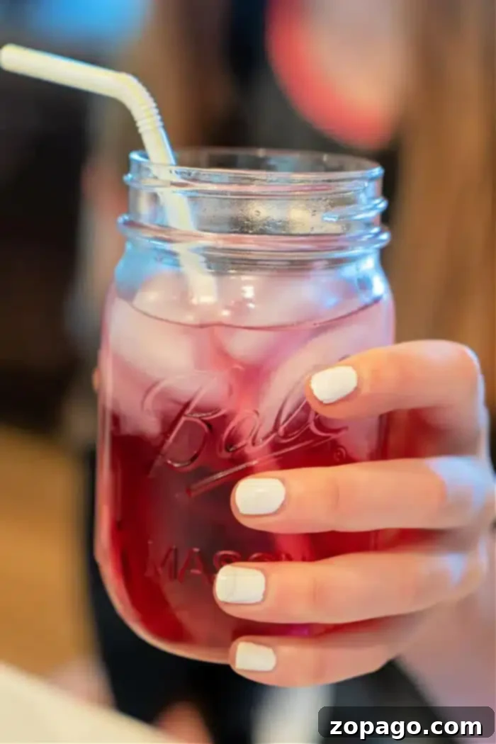 Whip Up a Refreshing Lotus Drink 2 A hand with white-painted nails holds a mason jar filled with a pink iced Lotus drink and a white straw. The jar is half-full, radiating refreshing vibes against the softly blurred background.