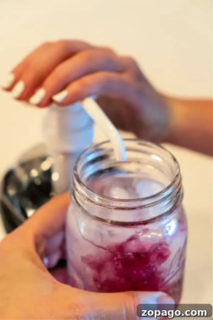 Whip Up a Refreshing Lotus Drink 6 A person pumps liquid from a dispenser into a mason jar filled with a refreshing Lotus iced drink. The person's other hand carefully holds the jar while the pump's nozzle hovers above it, adding the perfect touch. In the softly blurred background, hints of vibrant colors dance subtly out of focus.
