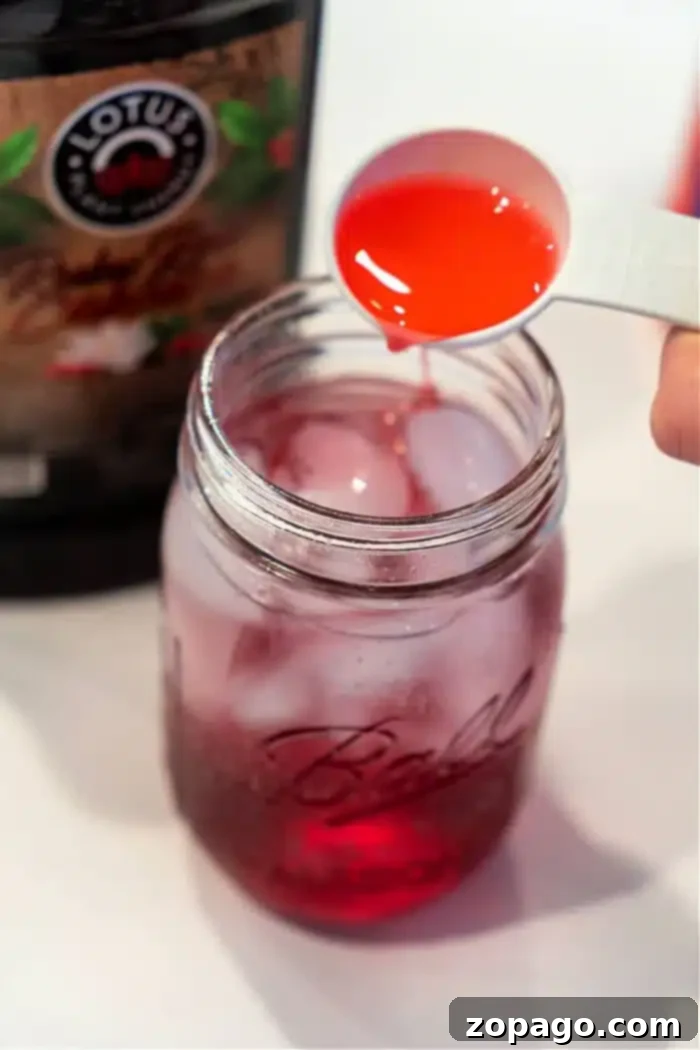 Whip Up a Refreshing Lotus Drink 7 A measuring spoon pours red liquid into a Ball Mason jar packed with ice and another red liquid, creating a tantalizing Lotus Drink. A partially visible bottle labeled Lotus hints at the refreshing flavors waiting to be discovered.