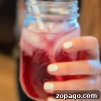 A hand holds a mason jar filled with a vibrant red Lotus drink, complete with ice cubes and a straw; nails are painted in crisp white.
