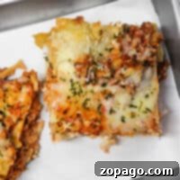 A steaming piece of lasagna topped with melted cheese and herbs rests on a metal tray, ready to delight your taste buds. Perfectly reheated in the oven, this classic dish offers a delectable experience.