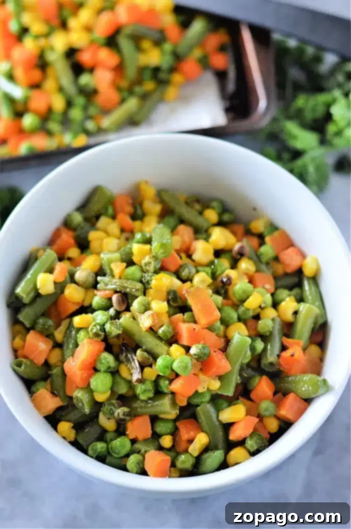 Crispy Roasted Frozen Vegetables 2 A bowl filled with roasted mixed vegetables, including green beans, corn, peas, and diced carrots. The vibrant colors suggest they were perfectly cooked from frozen. In the background, more vegetables await roasting on a tray.