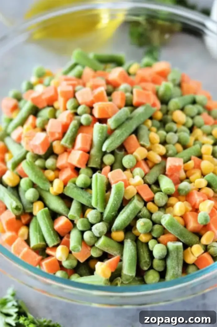 Crispy Roasted Frozen Vegetables 4 A glass bowl brimming with frozen mixed vegetables—green beans, peas, carrots, and corn—awaits the magic of roasting. The gray countertop sets a subtle stage with a hint of parsley for garnish.