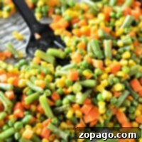 A close-up of roasting mixed vegetables, including green beans, corn, peas, and carrots on a baking sheet, with a black spoon resting on the side.