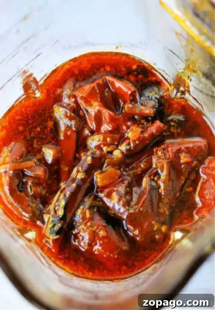 Birria Taco Sauce 2 A close-up of dried red chilies soaking in a rich, oily blend of spices and seasoning, reminiscent of a vibrant birria tacos sauce. The marinade is a luscious reddish-brown hue, with the chilies appearing softened and perfectly coated in the flavorful oil.