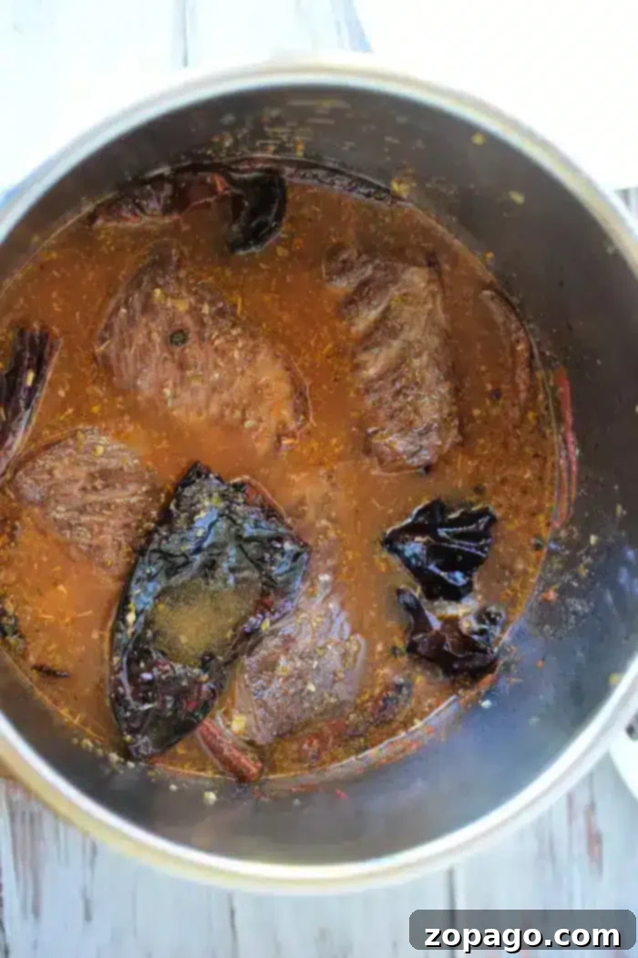 Birria Taco Sauce 7 A pot with chunks of beef simmering in a reddish-brown Birria sauce, accented by visible dried chili peppers and spices. The dish, rich in flavor and aroma, is likely being cooked on a stove.