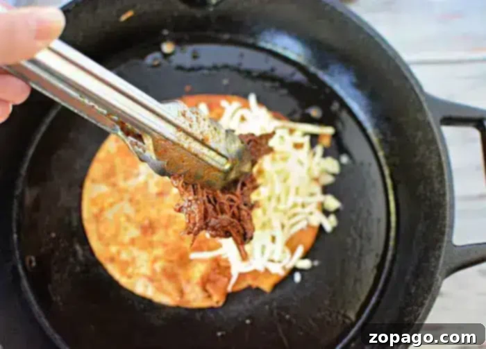 Birria Taco Sauce 8 A hand holds metal tongs, placing shredded beef onto a tortilla with melted cheese in a black skillet. The tortilla appears partially folded, suggesting the preparation of a quesadilla, while a drizzle of savory birria tacos sauce enhances its flavor.
