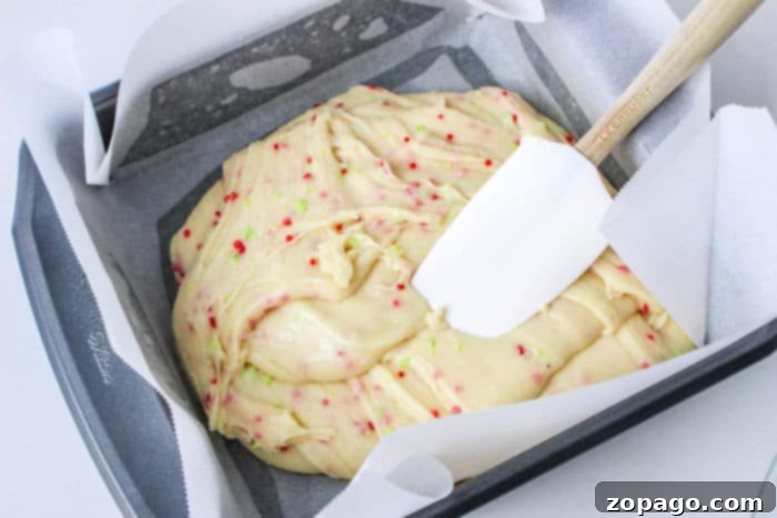 Cookie batter with colorful sprinkles in a parchment-lined pan, being smoothed with a spatula, brings to mind the richness of cookie mix fudge made with condensed milk.