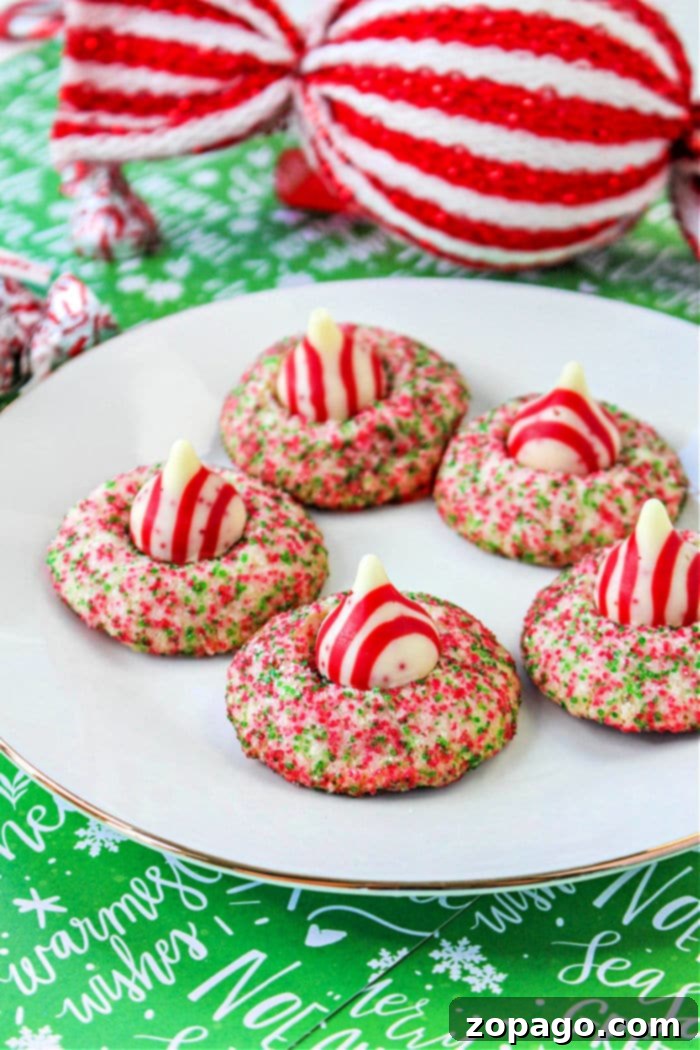 Artisan Blossom Cookies 8 A plate with five holiday-themed cookies topped with red and white striped candies, reminiscent of Hershey Kiss Christmas Cookies. The cookies are coated in vibrant red and green sprinkles. In the background, a large, wrapped decorative item boasts festive red and white stripes on a jolly green surface.
