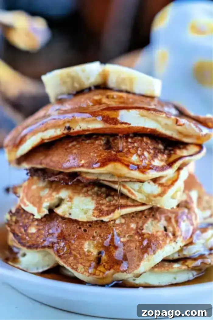 Banana Chocolate Chip Pancake Bliss 2 A close-up image of a stack of banana chocolate chip pancakes on a white plate. The pancakes are drizzled with syrup and topped with a slice of butter. The background is blurred, focusing attention on the delectable pancakes.