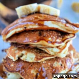 A stack of banana chocolate chip pancakes topped with banana slices and drizzled with syrup on a white plate.