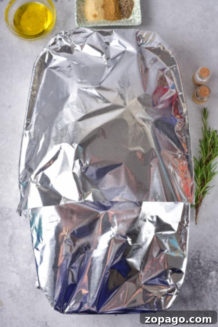 From Freezer to Fork: Cooking Delicious Duck 4 A frozen duck inside a baking dish, which is covered with aluminum foil and centered on the marble countertop. Nearby are a small bowl of olive oil, a plate with seasoning, two small shakers, and a sprig of rosemary, hinting at the flavorful preparation ahead.