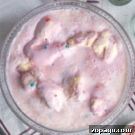A bowl of pink ice cream punch, inspired by the ultimate Pink Punch Recipe, is adorned with colorful sprinkles on a wooden table. Beside it lies a striped napkin and silver spoons, ready for indulgence.