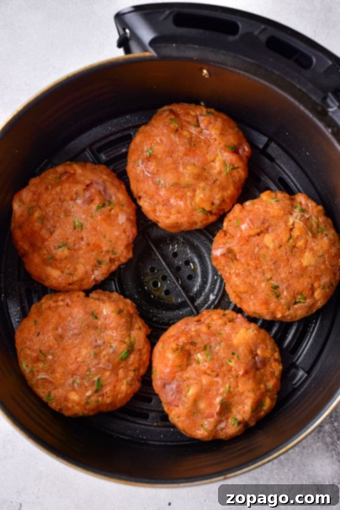 Five raw salmon patties seasoned with herbs sit in an air fryer basket, ready for a modern twist on cooking. These aren't your Old Fashioned Salmon Patties, but they're sure to awaken nostalgic flavors with every bite.