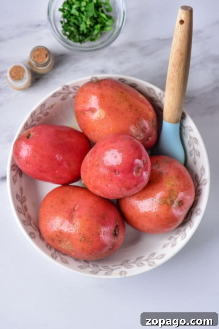 Perfectly Roasted Red Potatoes 3 A white bowl with a floral pattern contains five red potatoes and a light blue vegetable peeler with a wooden handle, perfect for prepping baked red potatoes. In the background, on a marble surface, there's a small bowl of chopped herbs and two spice jars.
