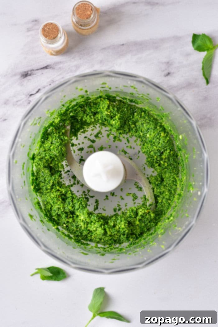 Quick & Easy Pine Nut Pesto 4 Green pesto without pine nuts swirls in a food processor bowl on a marble countertop, accompanied by fresh basil leaves and spice jars nearby.