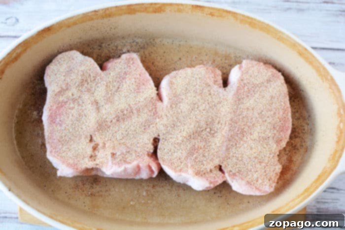 Perfect Pork Chops from Frozen 5 Three uncooked, seasoned frozen pork chops sit in an oval ceramic baking dish on a wooden surface, ready to be transformed into a delicious meal when baked in the oven.