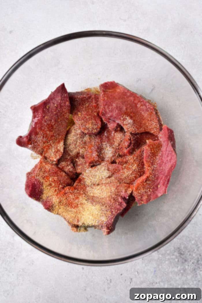 Mastering Beef Heart Cooking 4 A glass bowl containing raw beef heart pieces all coated with various seasonings, is placed on a light gray surface.