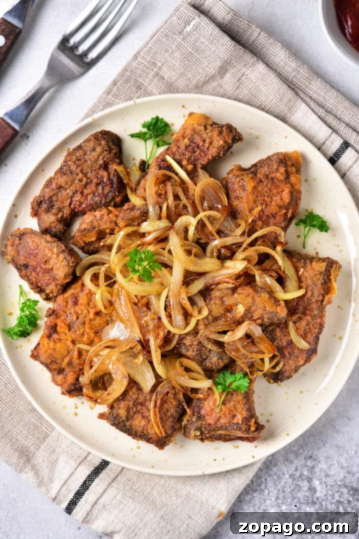 Mastering Beef Heart Cooking 7 A plate of beef heart slices and onions garnished with fresh parsley features browned liver pieces alongside tender beef heart slices, all topped with sautéed onions. The dish rests on a cloth napkin with a fork and knife beside it.