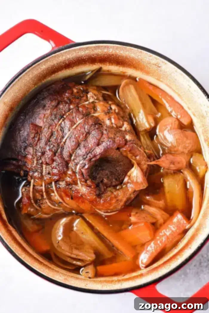Slow Roasted Beef Shoulder 5 A generously cooked beef roast shoulder, securely tied with kitchen string, rests nestled in a red Dutch oven. It's surrounded by vibrant chunks of carrots, celery, and onions, all bathing in a savory brown broth infused with visible spices. This wholesome image captures the essence of a slow-cooked, tender meal.
