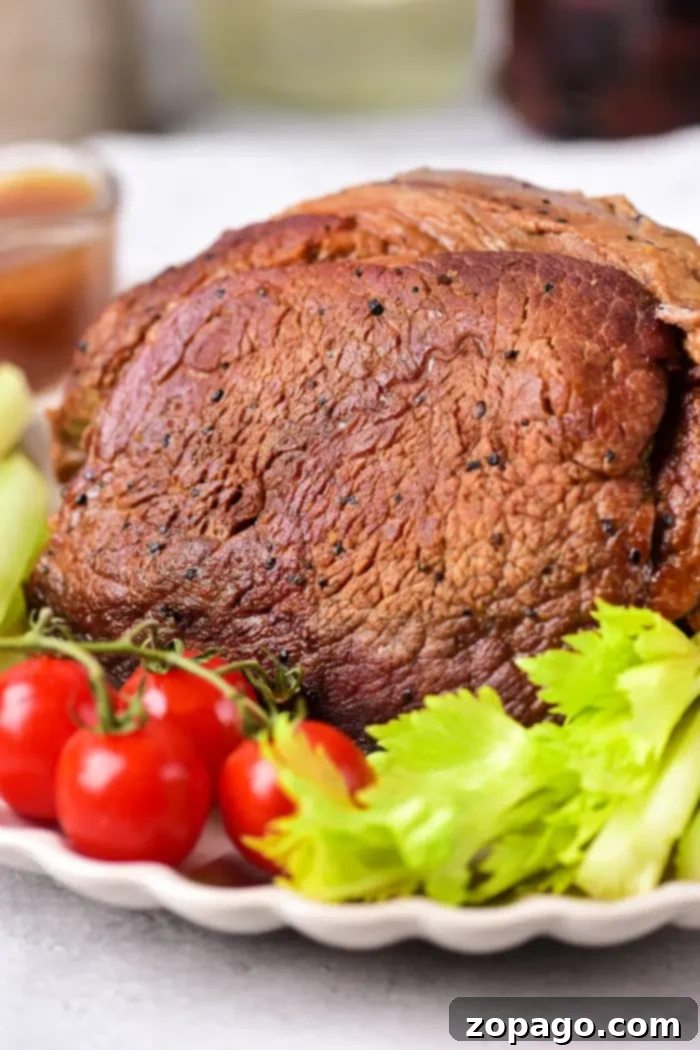 Slow Roasted Beef Shoulder 7 A roasted beef roast shoulder served on a white plate, garnished with fresh cherry tomatoes and celery stalks. The beef appears well-seasoned with a brown crust. A blurred bowl of sauce is visible in the background.