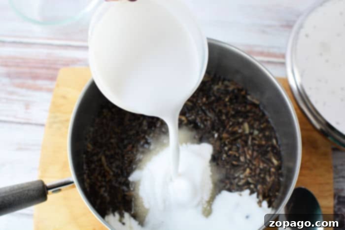Pouring creamy coconut milk into a pot filled with black rice and sugar on a wooden surface, creating a luscious black rice pudding.
