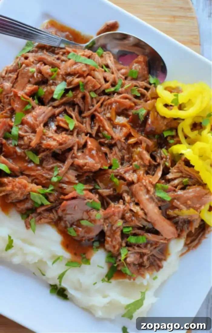 Slow-Cooked Mississippi Pork 2 A plate of perfectly shredded Mississippi Pork Roast, served generously over creamy mashed potatoes and garnished with fresh chopped parsley. A side of vibrant yellow pickled peppers complements the dish, with a spoon resting invitingly on the plate.