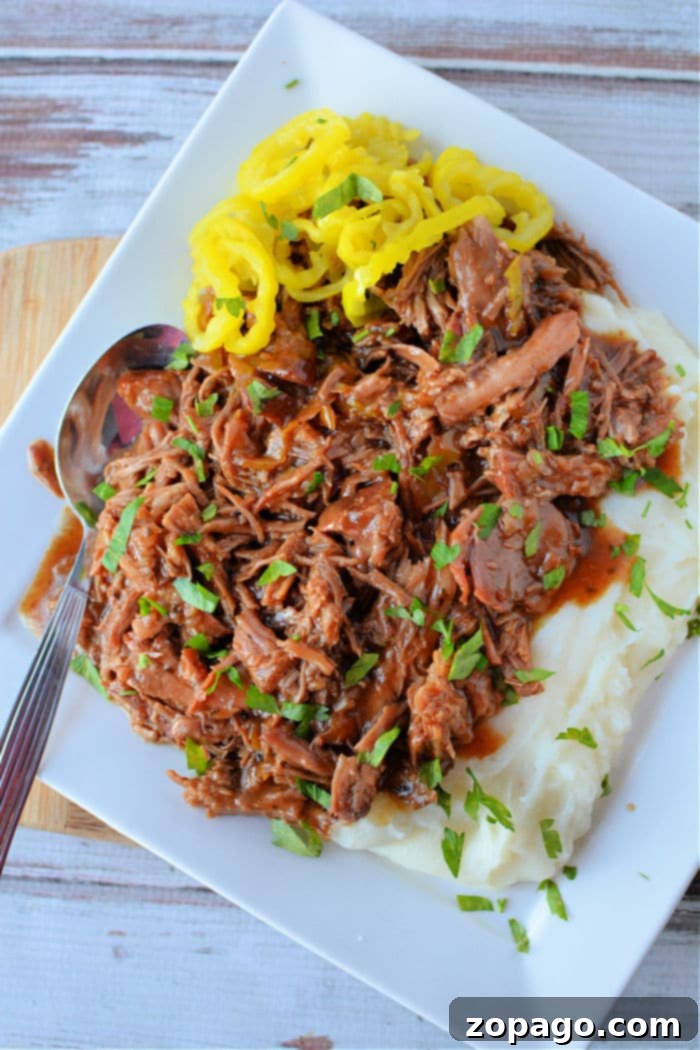 Slow-Cooked Mississippi Pork 6 A white rectangular plate showcases succulent shredded pork bathed in its rich gravy, garnished with fresh herbs, alongside a mound of creamy mashed potatoes. Bright yellow pepper slices add a vibrant touch, with a metal spoon resting on the plate's edge.