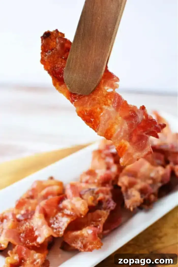 Perfect Oven Bacon on Foil 7 A piece of crispy bacon held by wooden tongs above a white rectangular plate filled with more bacon strips. The background is blurred, and the focus is on the bacon being lifted, showcasing perfectly cooked strips—just like what you'd get if you knew how to cook bacon in the oven with aluminum foil